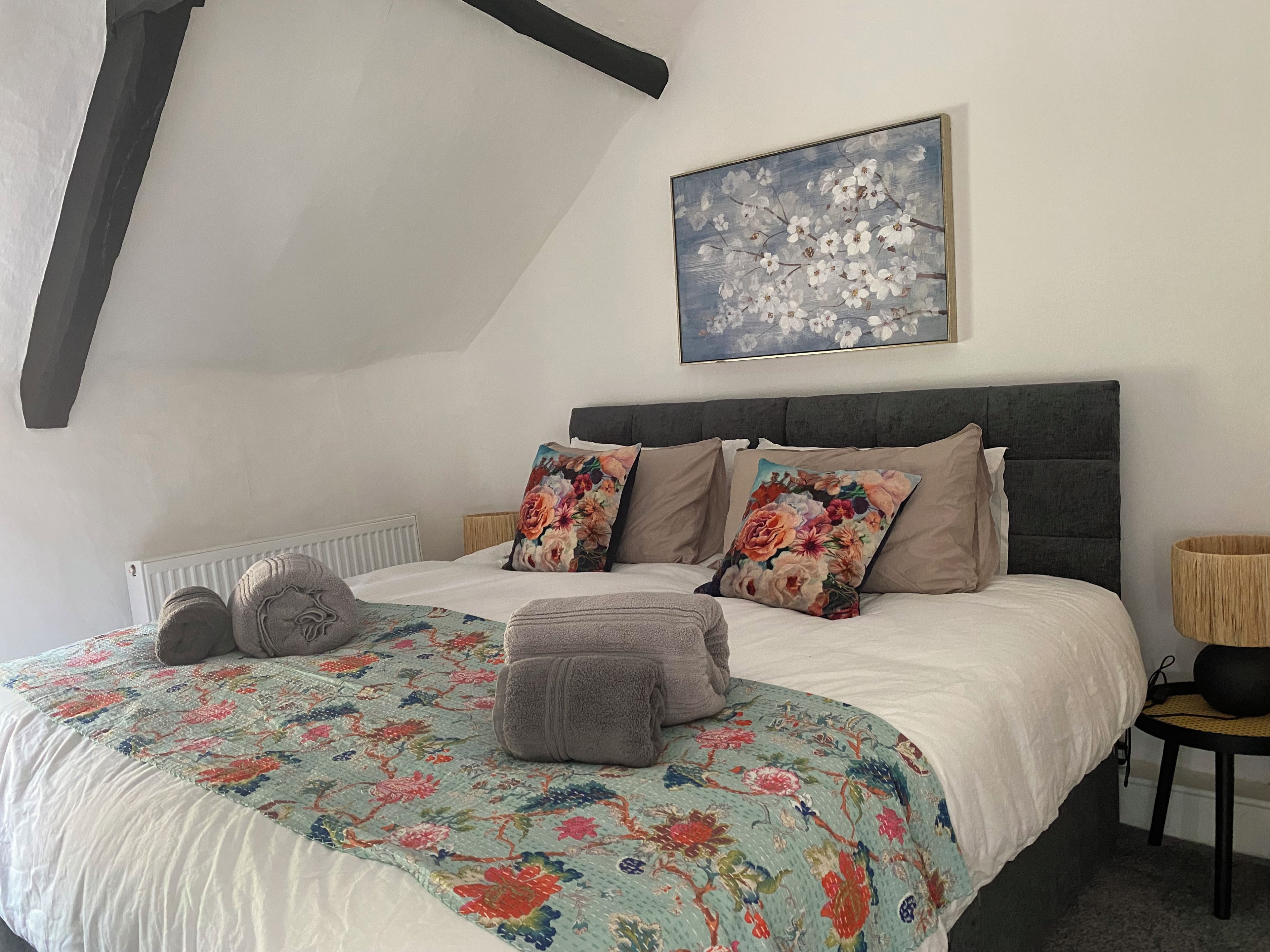 Un dormitorio con cama, almohadas, colcha, mesita de noche, lámpara y un cuadro en la pared.
