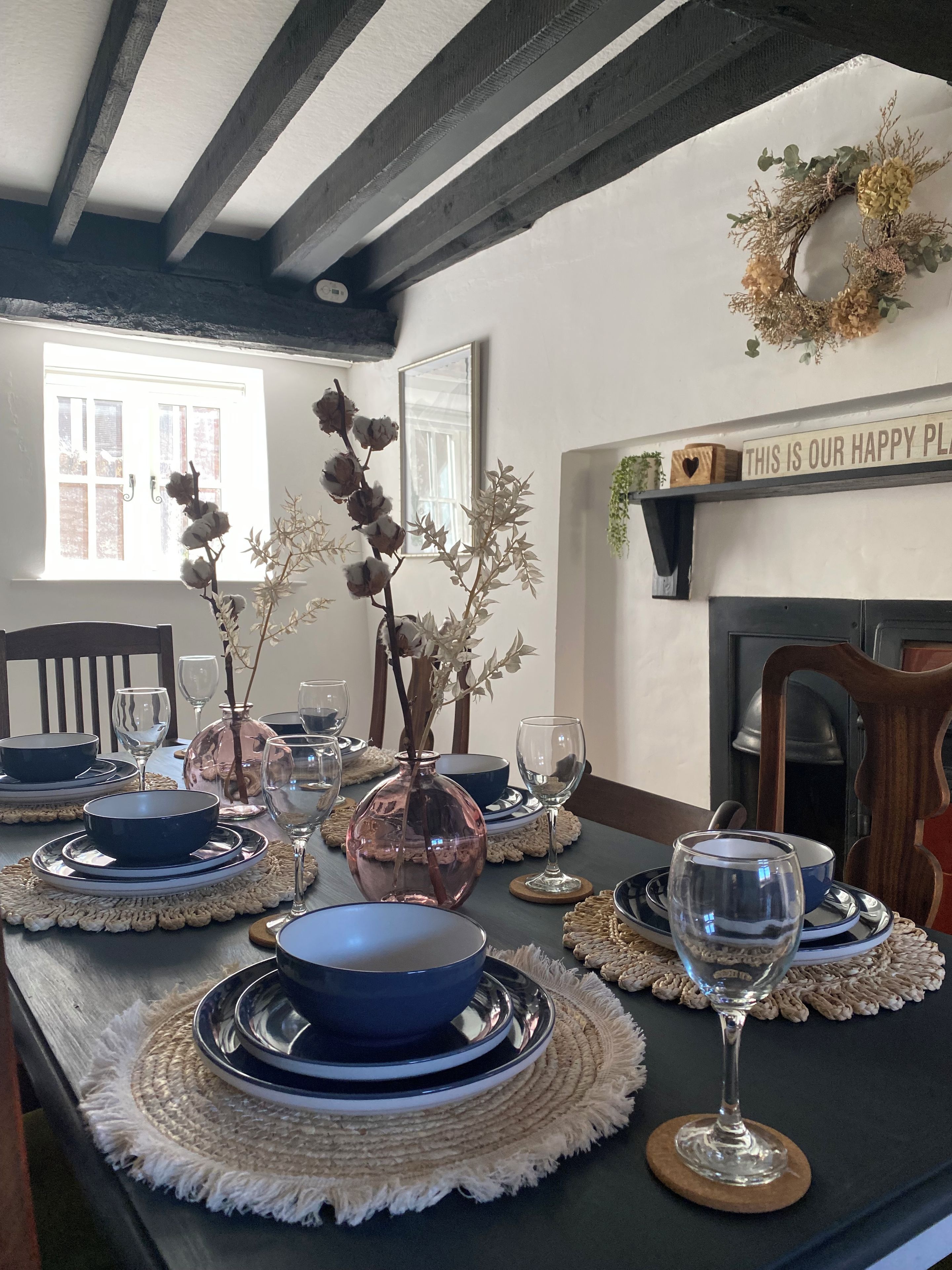 A dining table set with blue plates, silverware, and glassware, with a vase of dried flowers and a decorative wreath on the wall.