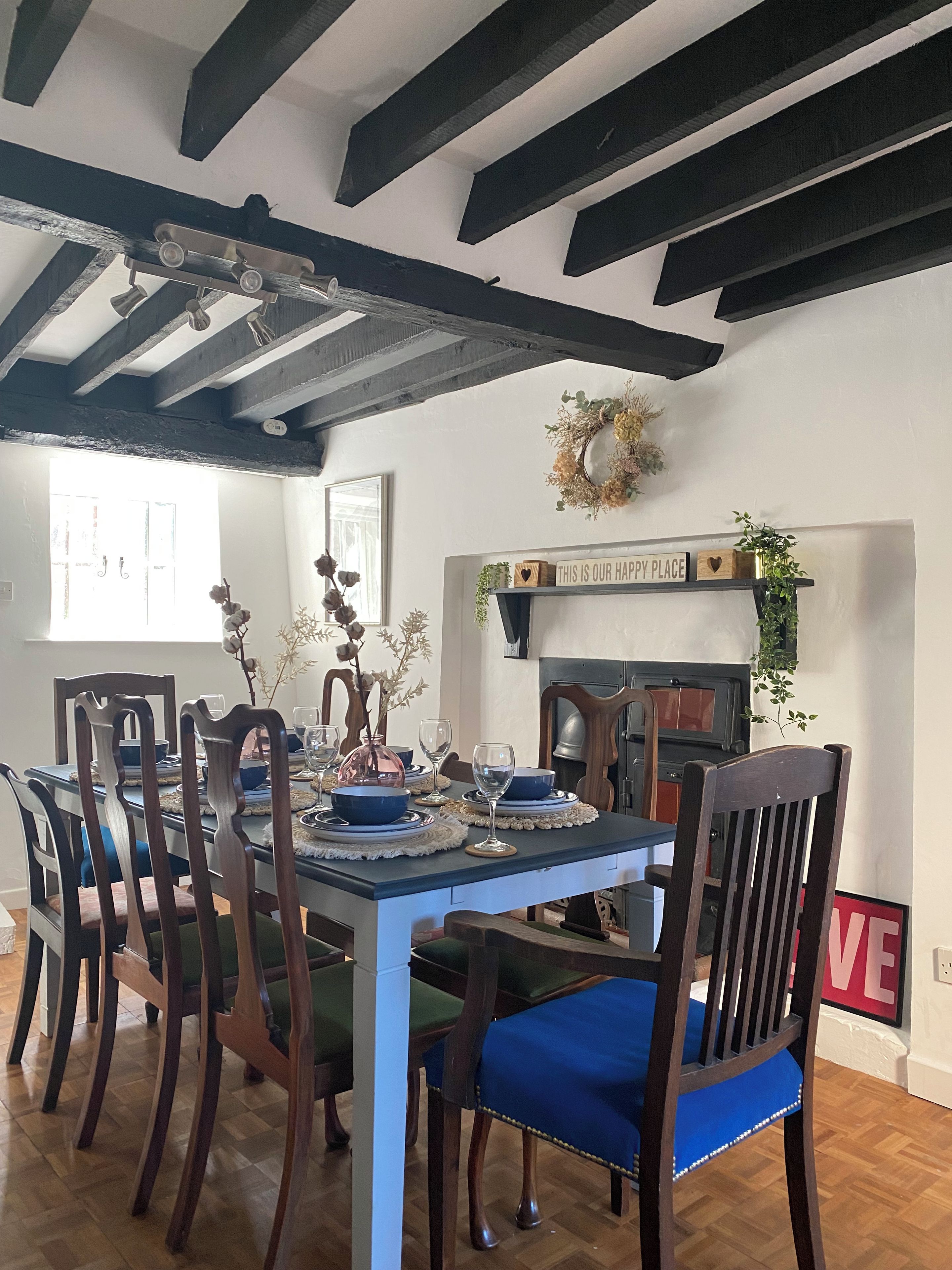 Uma sala de jantar com mesa de madeira posta para quatro pessoas, rodeada por cadeiras com almofadas azuis. Uma lareira com uma placa decorativa acima é visível ao fundo.