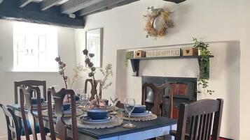 Uma sala de jantar com mesa de madeira posta para quatro pessoas, rodeada por cadeiras com almofadas azuis. Uma lareira com uma placa decorativa acima é visível ao fundo.