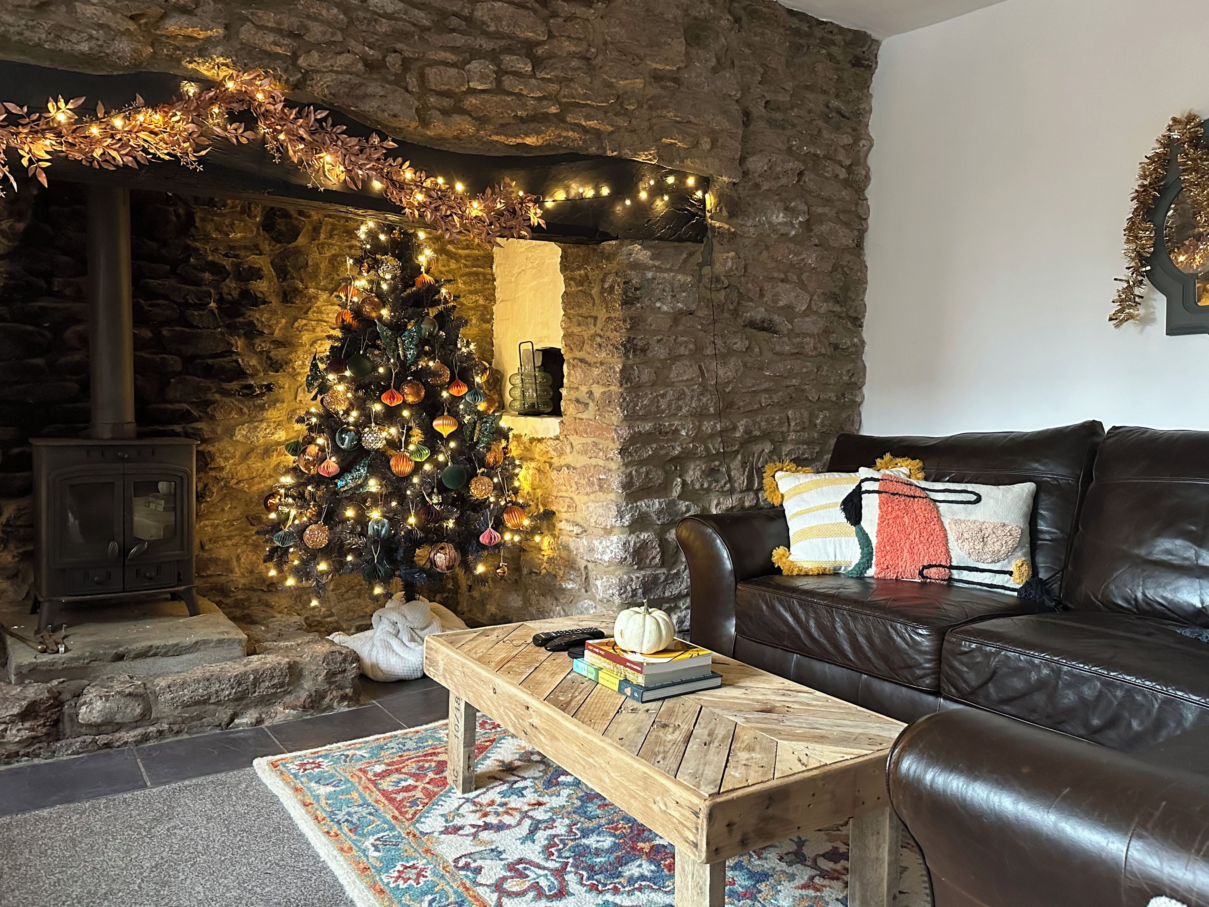 A cozy living room with a stone wall, a fireplace, a Christmas tree, a leather sofa, and a coffee table with books and a pumpkin.