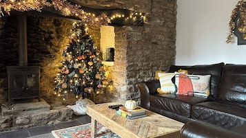 A cozy living room with a stone wall, a fireplace, a Christmas tree, a leather sofa, and a coffee table with books and a pumpkin.