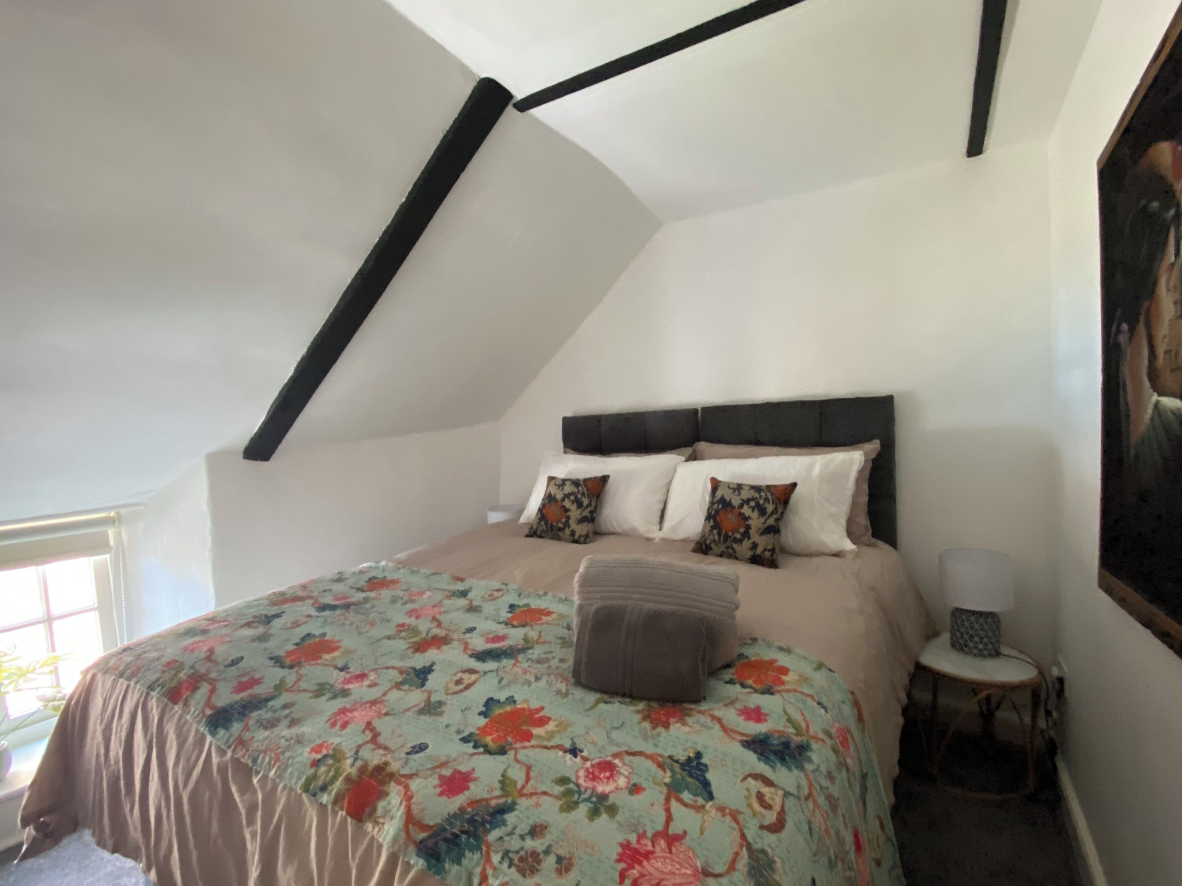 Ein Schlafzimmer mit schrägverdeckter Decke, einem Bett mit floralem Bettbezug, einem Nachttisch und einer Lampe.