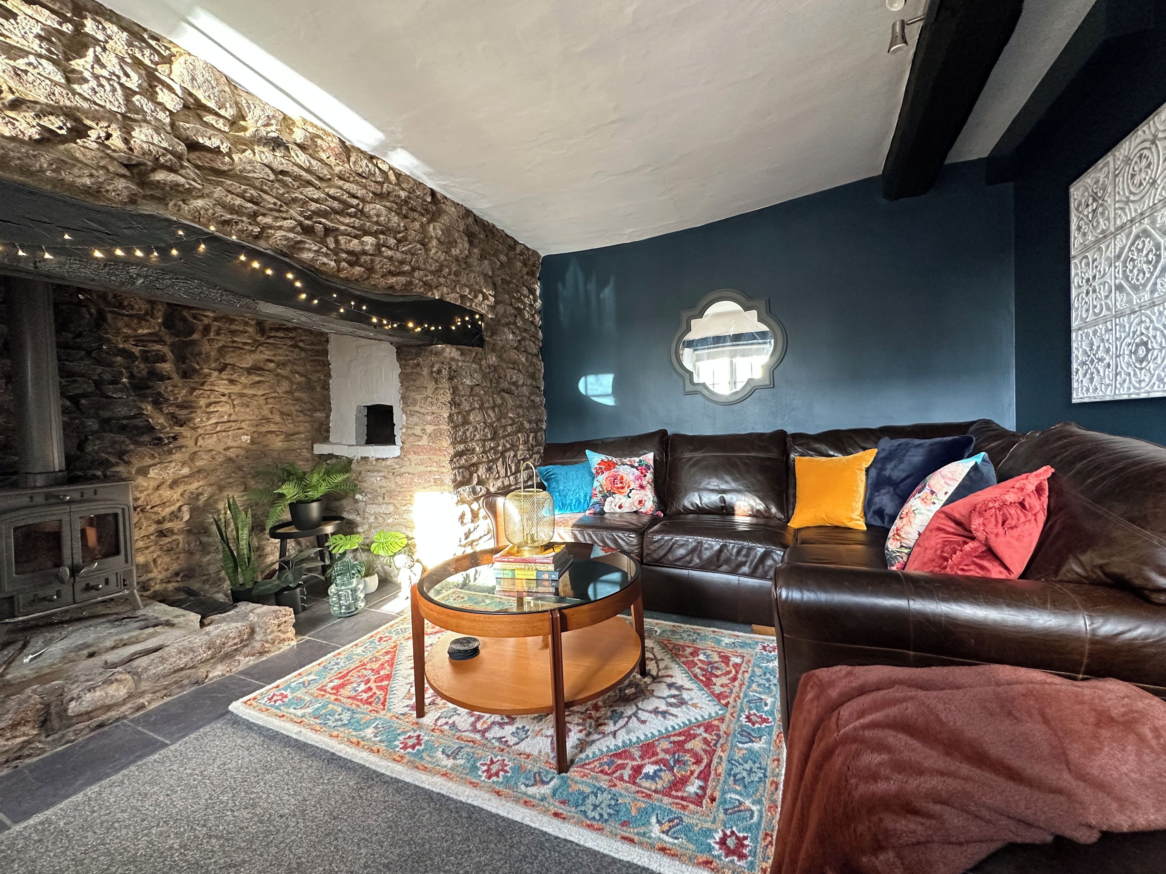 A cozy living room with a stone wall, a fireplace, a leather sofa with colorful cushions, and a patterned rug.