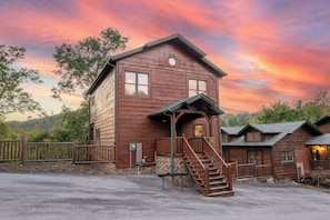 A wooden lodge with a porch and stairs leading to the entrance, surrounded by a stone wall and a fence. - Bear Cave | Theater, Arcade & Hot Tub | 3 King Suites | Sleeps 10 (Gatlinburg)