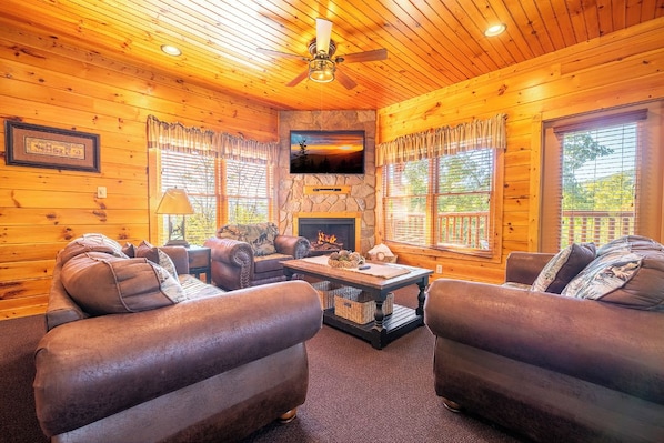 A log cabin interior with a fireplace, leather sofas, a television, and a ceiling fan. - Bear Cave | Theater, Arcade & Hot Tub | 3 King Suites | Sleeps 10 (Gatlinburg)