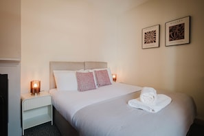 A hotel room with a bed, pillows, a rolled towel, two wall-mounted abstract art pieces, and bedside lamps. - Comfortable 3 Bedroom Townhouse Sleeps 6 (Northampton)