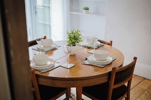 A round wooden dining table with white plates, bowls, and wine glasses set for four. There is a small potted plant on the table, and the room has white walls and a wooden floor. - Comfortable 3 Bedroom Townhouse Sleeps 6 (Northampton)