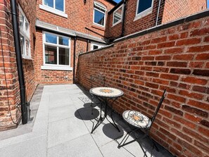 Een smal terras buiten met twee ronde tafels en stoelen, bakstenen muren en een heldere lucht.