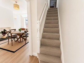 Een trap met beklede, beige treden en witte spijlen, die leidt naar een ruimte met een houten vloer en een eethoek met een ronde tafel en stoelen.