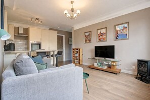 A modern living room with a sofa, a TV on a glass stand, and a kitchen in the background. - Greenfields Apartment (Christleton)