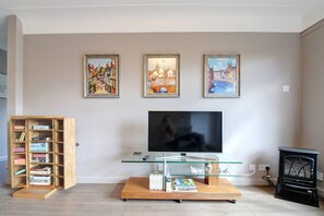 A living room with a flat-screen TV, a wooden bookshelf, and three framed paintings on the wall. - Greenfields Apartment (Christleton)