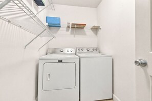 Ein Waschraum mit zwei weißen Waschmaschinen und Trocknern, einem Metallregal mit Wäschekörben und einem Lüftungsgitter an der Wand.