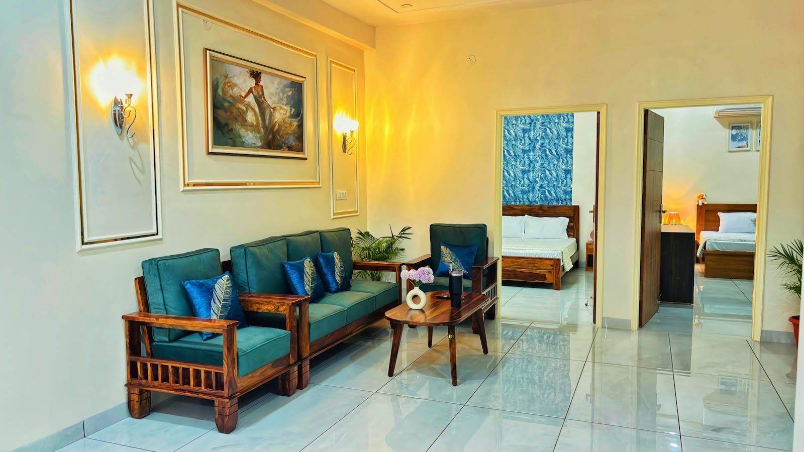 A hotel room with a sofa set, a wooden coffee table, a bed with a blue patterned headboard, and a wall-mounted light fixture.