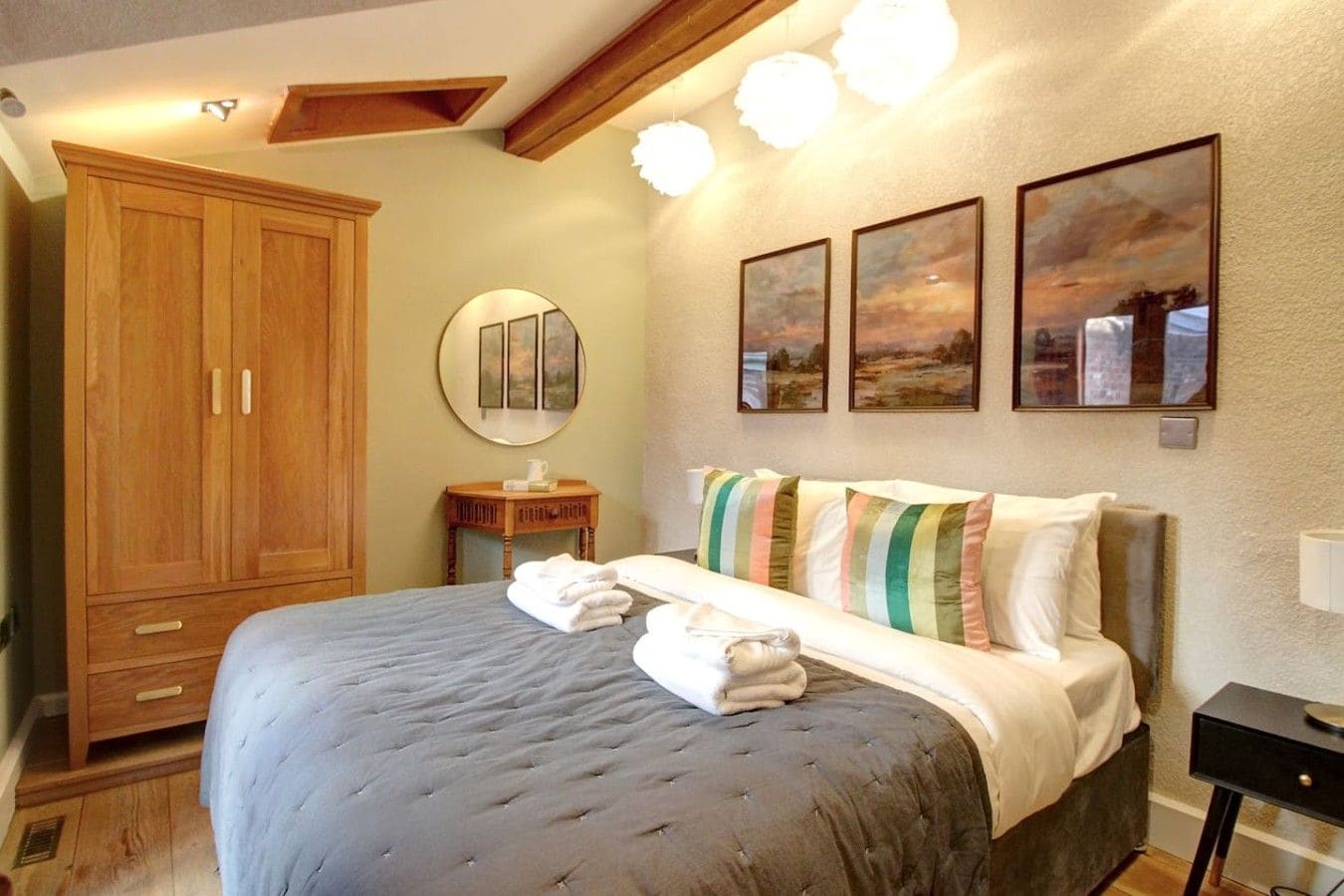 Una camera da letto con un letto grande, un armadio in legno, uno specchio rotondo e tre quadri raffiguranti paesaggi.