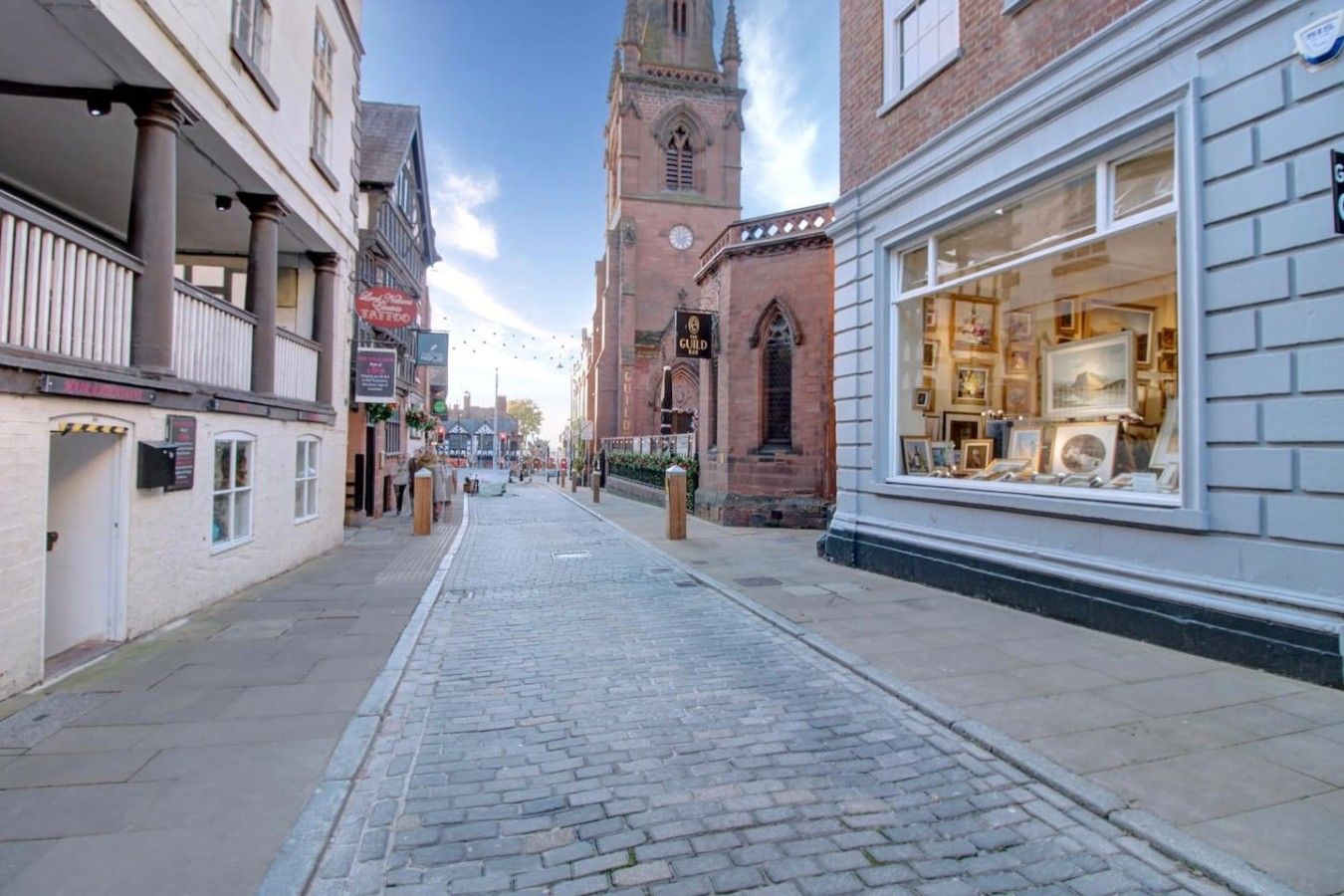 Una calle empedrada con edificios históricos, una torre de iglesia y una tienda con cuadros enmarcados.