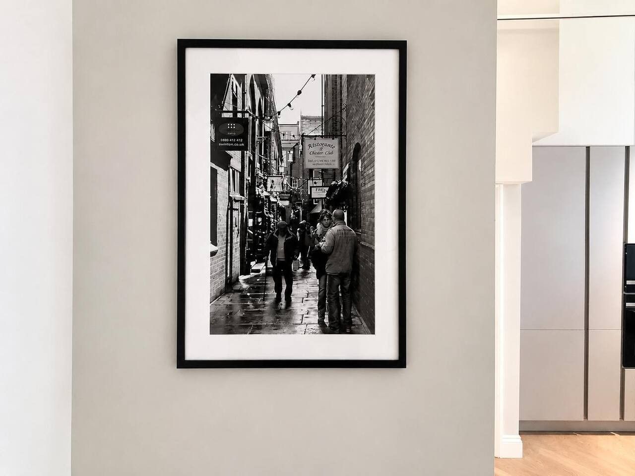 A framed black and white photograph of a narrow street with people walking, a restaurant sign, and a building with a sign that reads 'Free.'