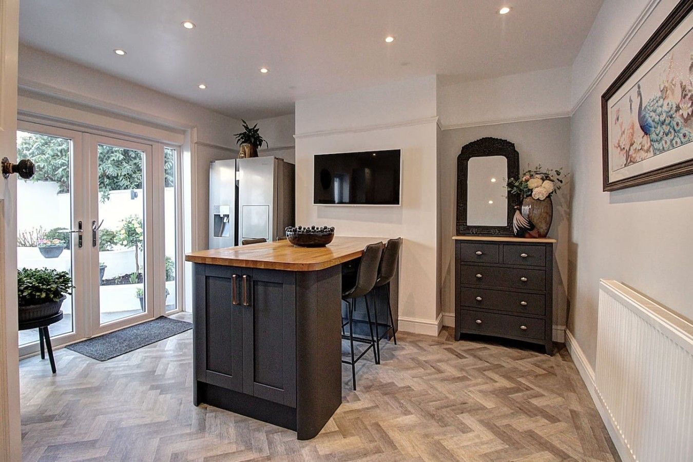 A kitchen with a central island, a dining area with chairs, a refrigerator, a flat-screen TV, and a decorative mirror.