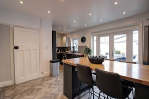 A modern kitchen with a central island, a dining area with chairs, and a view through large windows. - The Botanical Holiday by Cheshire Escapes (Cheshire West and Chester)