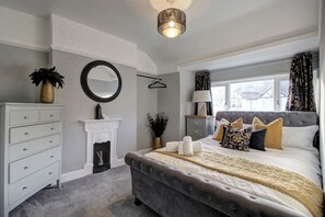 A bedroom with a large bed, a dresser, a mirror, a fireplace, and a window with curtains. - The Botanical Holiday by Cheshire Escapes (Cheshire West and Chester)