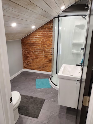 Un baño con un lavamanos blanco, cabina de ducha con mampara de vidrio, inodoro y un tapete azul en el suelo.