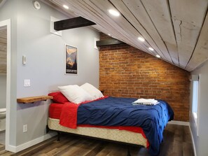 A bedroom with a bed, a wooden shelf, a brick wall, and a poster on the wall. - Lovely renovated one bedroom getaway (Meaford)
