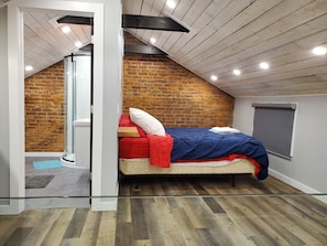 Un dormitorio con techo inclinado, pared de ladrillo, cama con ropa de cama roja y azul, y ventana con persiana.