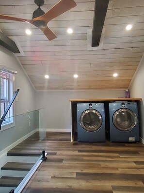 A laundry room with two front-loading washing machines, wooden flooring, a glass railing, and a ceiling fan. - Lovely renovated one bedroom getaway (Meaford)