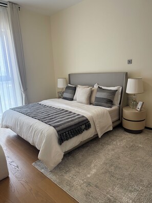 A neatly made bed with a grey headboard, a bedside table with a lamp, and a window with curtains. - Elegant 2-Bedroom Luxury Retreat at GTC Residency (Nairobi)