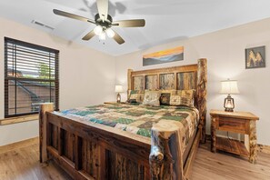 A bedroom with a wooden bed, a ceiling fan, a window with blinds, a painting, and a lamp on a wooden nightstand. - 3BD 3BA Cabin Private Hot Tub Indoor/Outdoor Pools Lazy River! (Hollister)