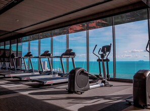A gym with treadmills, elliptical machines, and a rowing machine, featuring large windows with a view of the ocean and cityscape. - Luxury Condo With Panoramic Sea View Sky Pool (Pattaya)