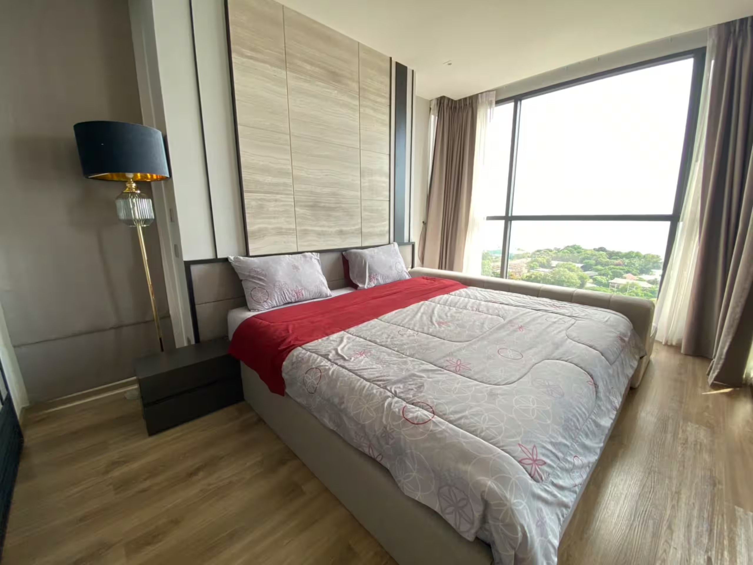 A modern bedroom with a large bed, a nightstand, a lamp, and a view of greenery through the window.