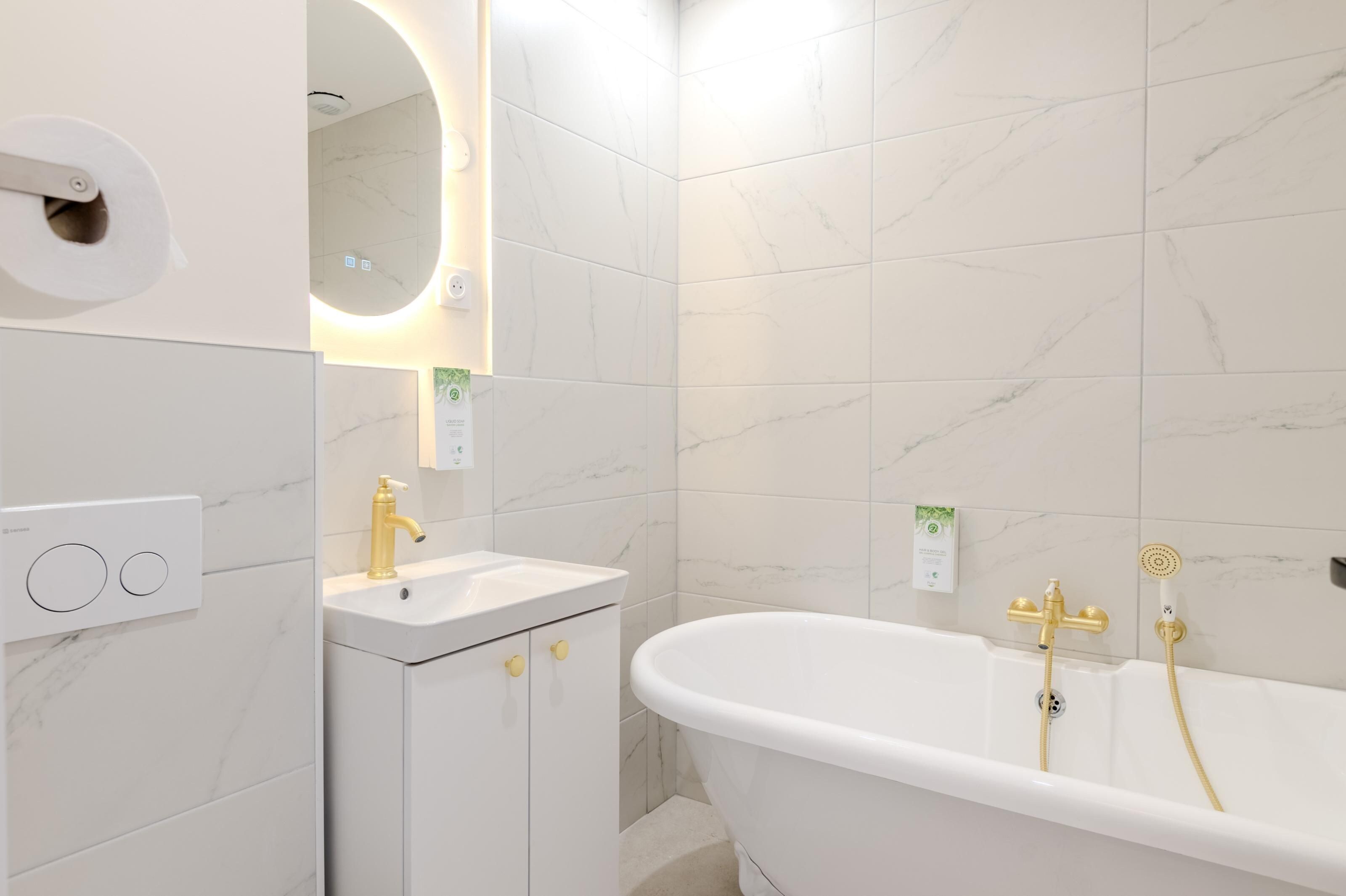 Un baño con una bañera blanca, un lavamanos con grifo dorado y un espejo con luz circular.