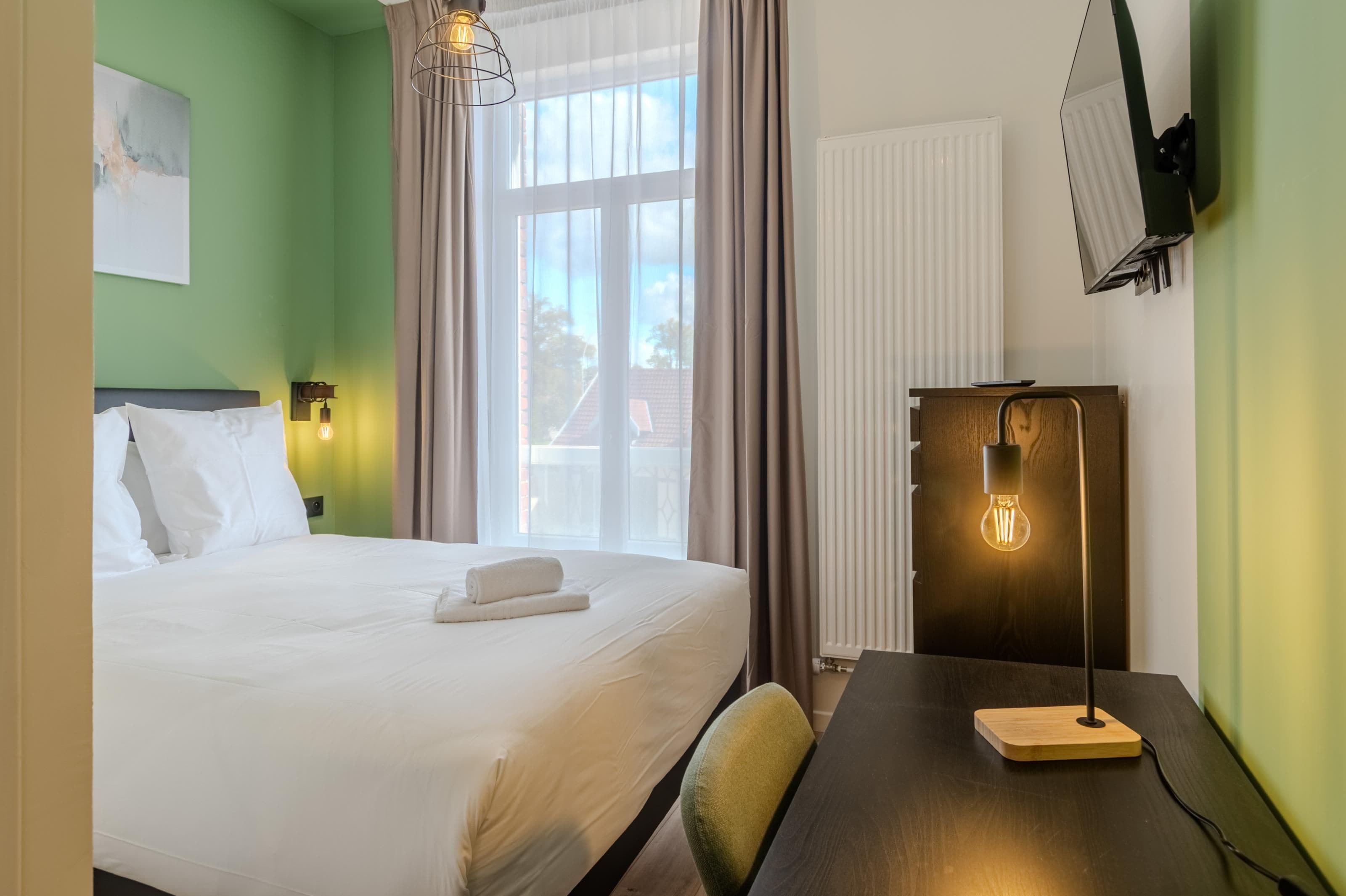 Una camera d'albergo con un letto, una scrivania, una sedia, una lampada da tavolo, una TV a parete e una finestra con tende.