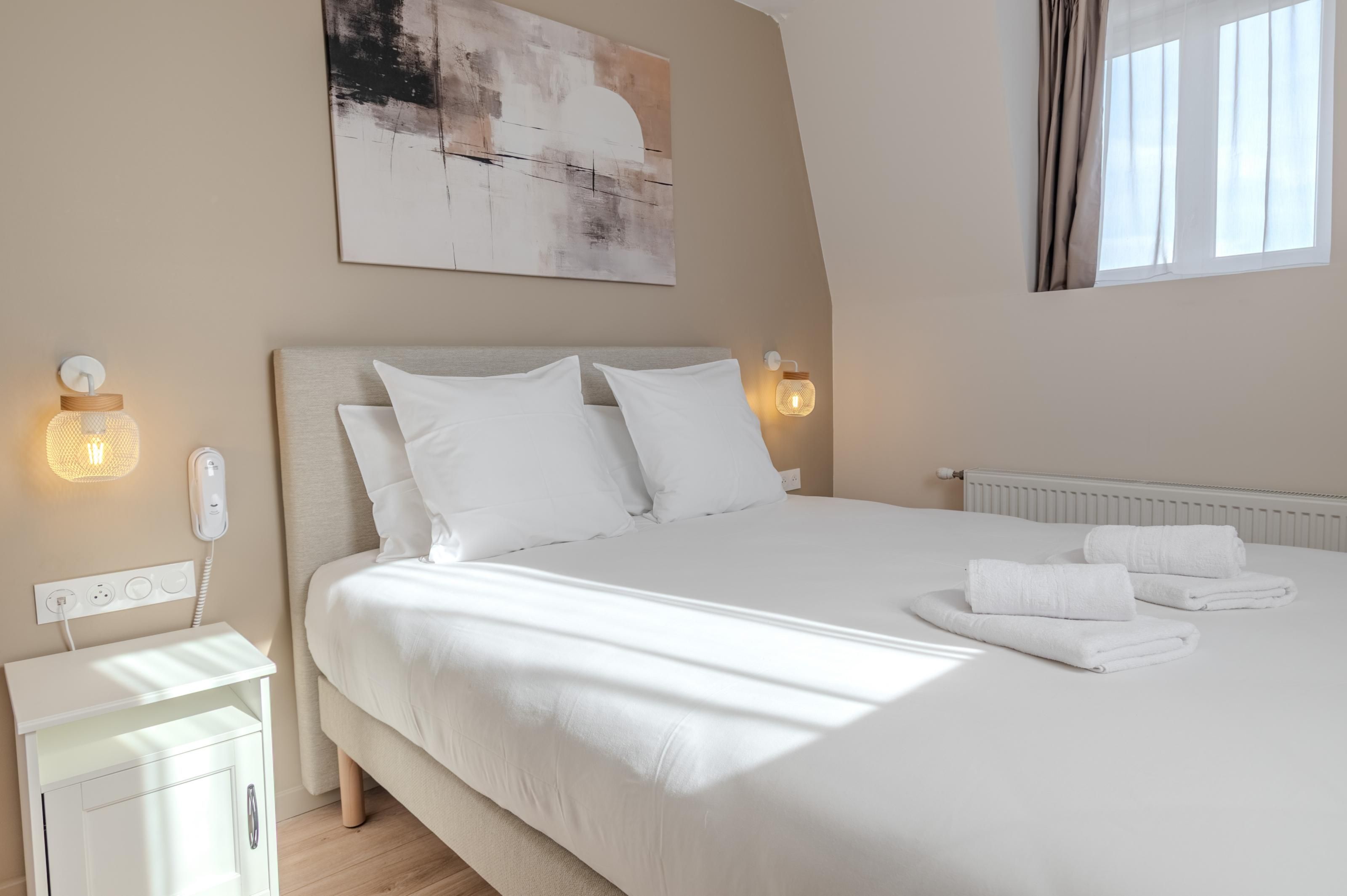 A neatly made bed with white linens and pillows, a bedside table with a lamp, and a wall-mounted light fixture.