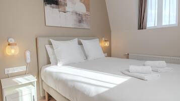 A neatly made bed with white linens and pillows, a bedside table with a lamp, and a wall-mounted light fixture.