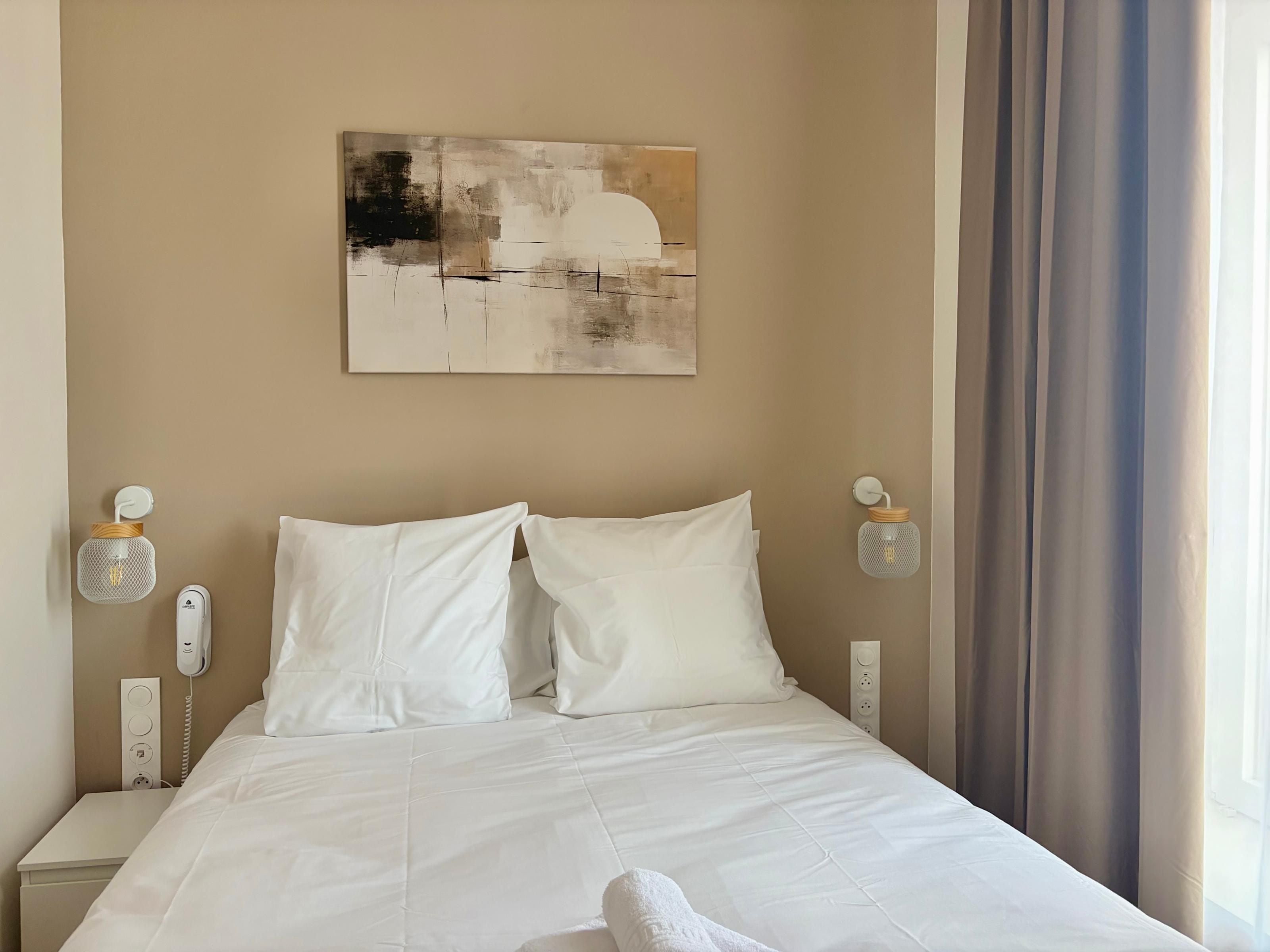 A hotel room with a bed, a painting on the wall, a telephone, and a lamp.