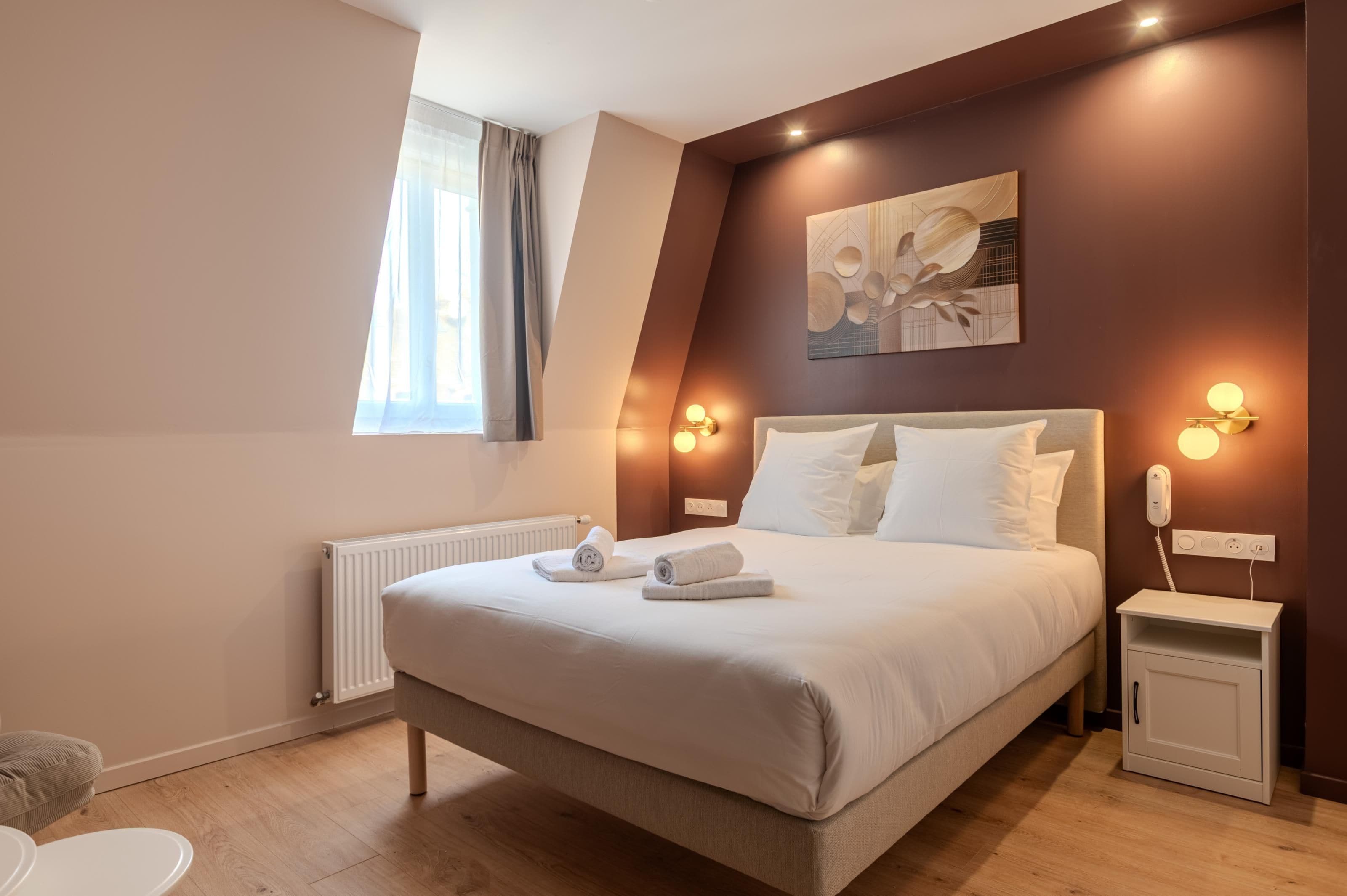 A hotel room with a large bed, bedside tables, a radiator, a window with curtains, and a wall-mounted artwork.