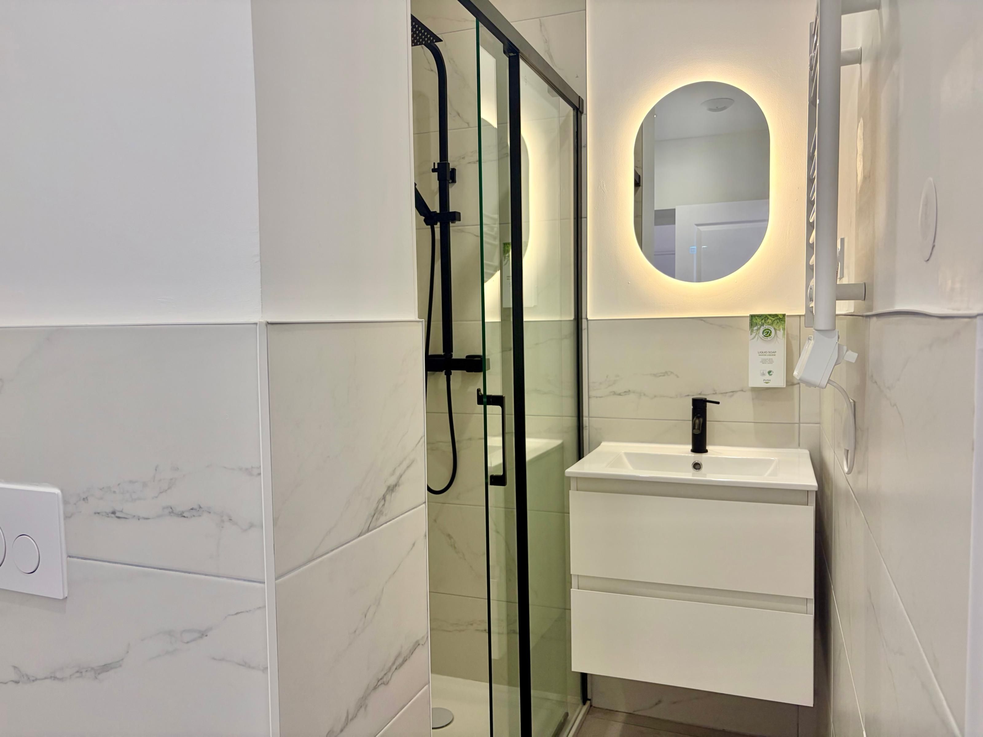 Een moderne badkamer met een douchecabine van glas, een wit wastafelmeubel met een zwarte kraan en een spiegel met ronde verlichting.
