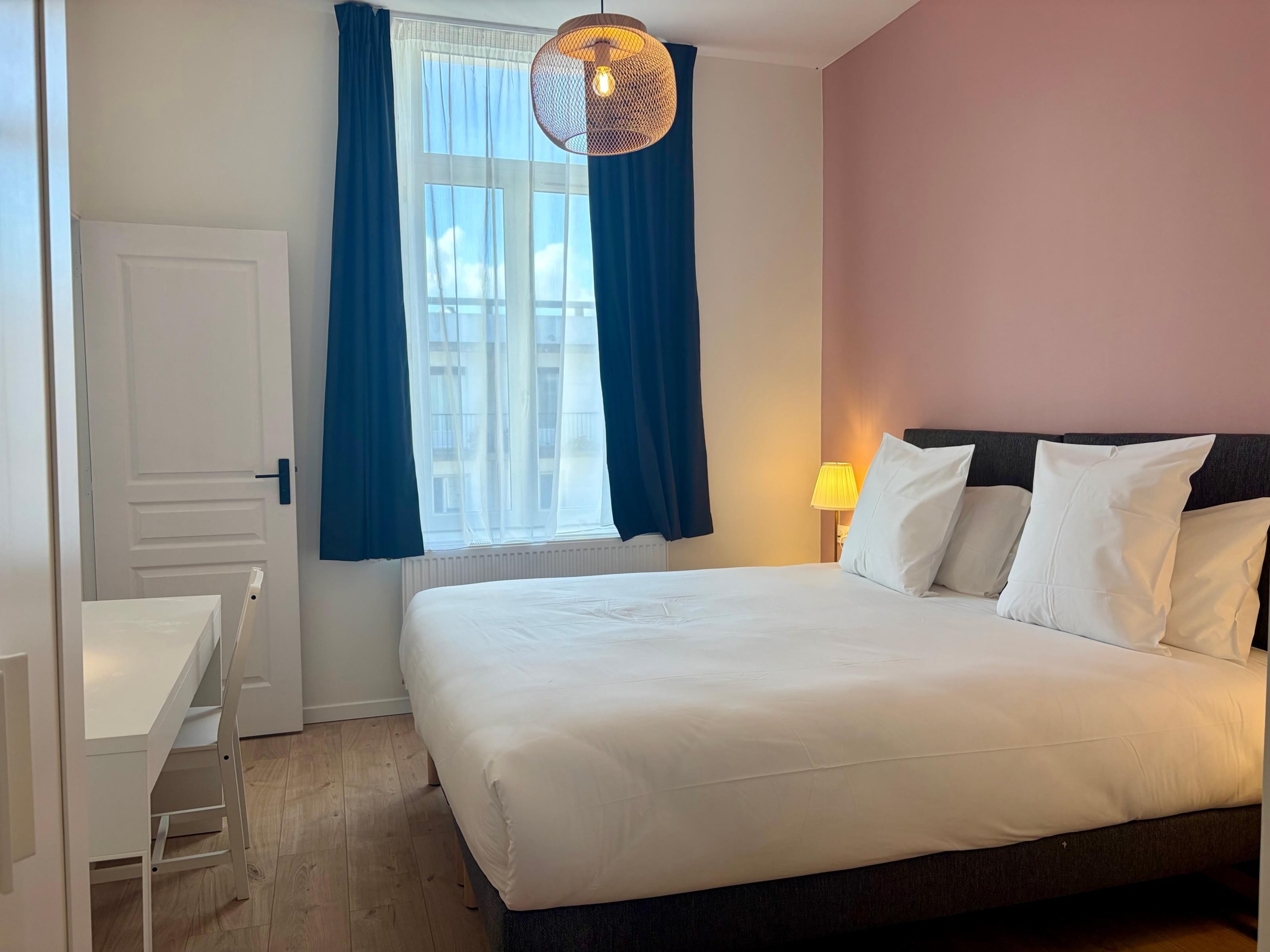 Hotellihuone, jossa on sänky, työpöytä, tuoli, ikkuna verhoilla ja katosta roikkuva valaisin.