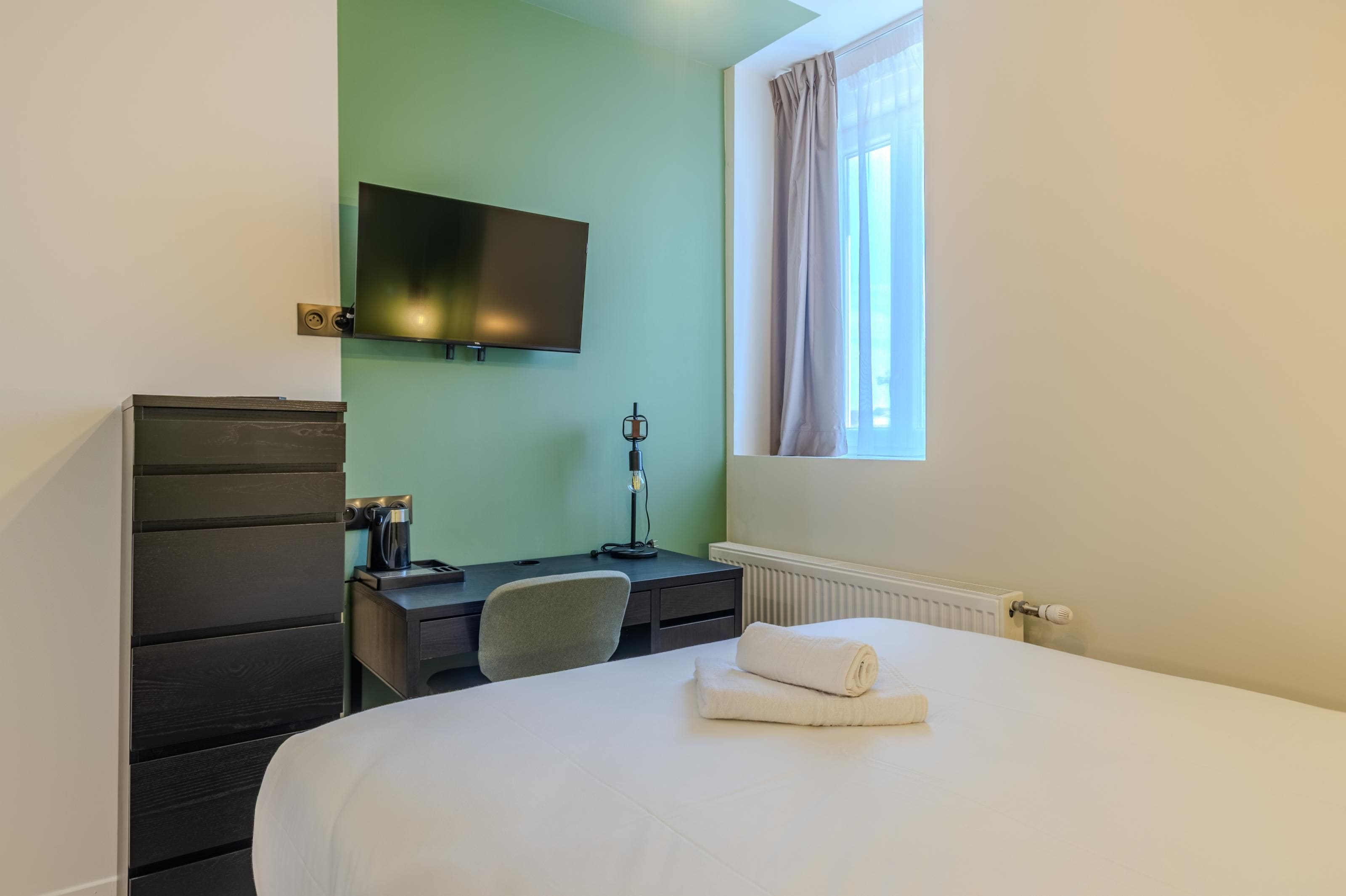 A hotel room with a bed, a desk, a chair, a television mounted on the wall, and a window with curtains.