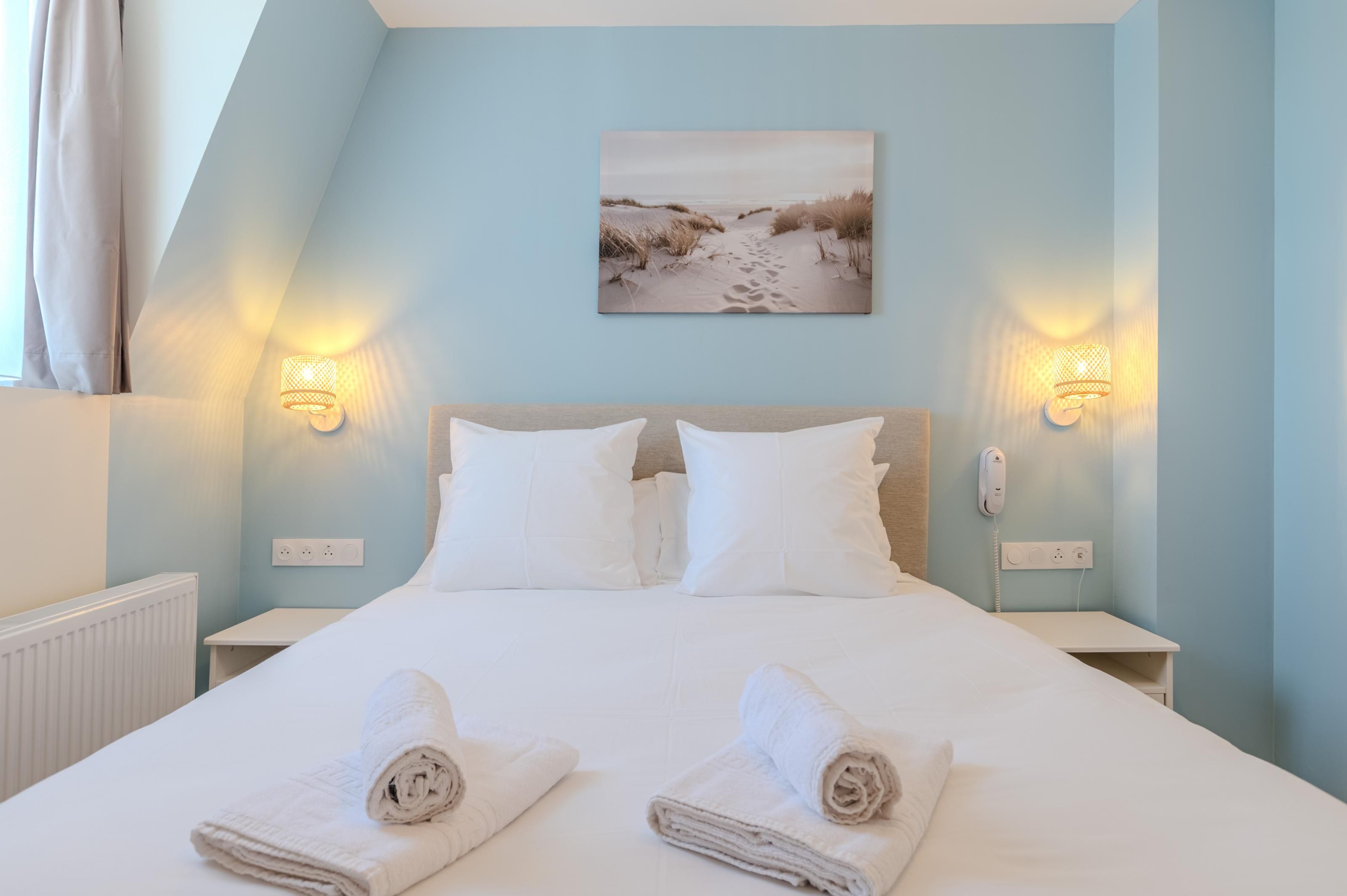 A bedroom with a bed, two bedside tables, a nightstand, a wall-mounted phone, and a painting of a beach.