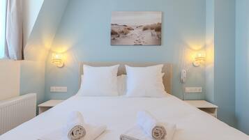 A bedroom with a bed, two bedside tables, a nightstand, a wall-mounted phone, and a painting of a beach.