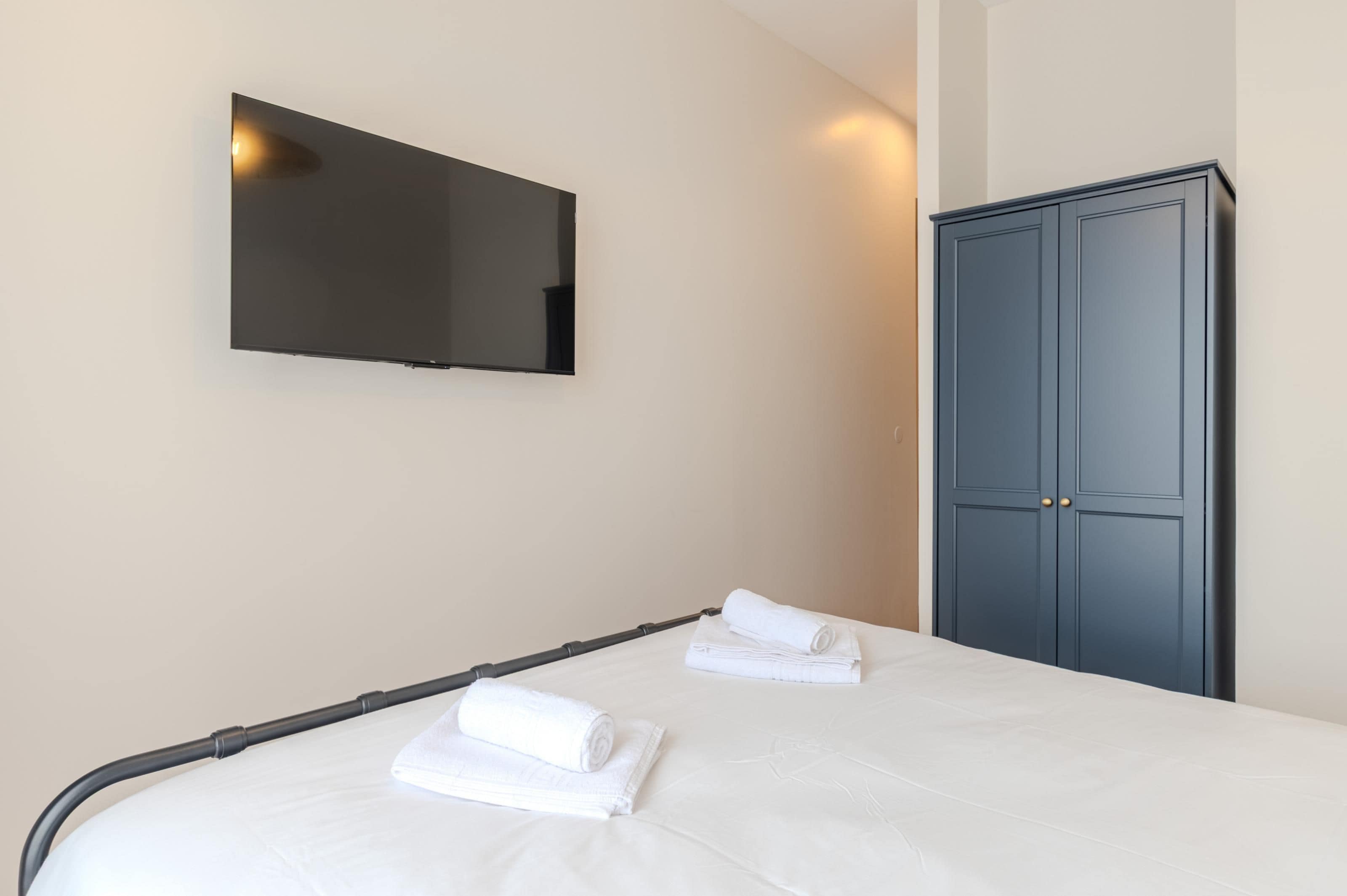 Une chambre d’hôtel avec un lit, deux serviettes enroulées, une armoire et un téléviseur fixé au mur.