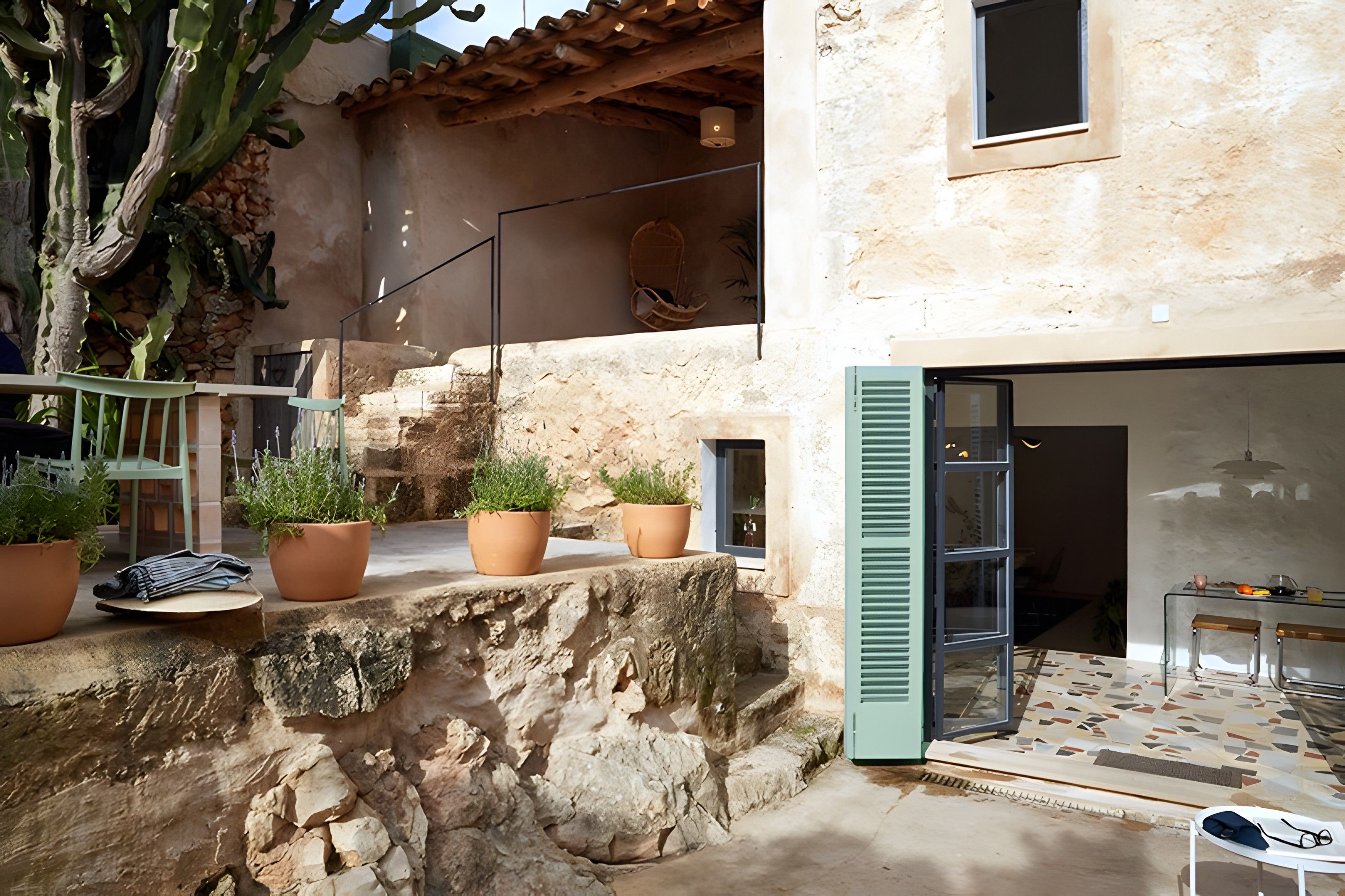 Un mur de pierre agrémenté de plantes en pot, un escalier et une porte vitrée donnant sur un espace intérieur doté d’un sol carrelé.
