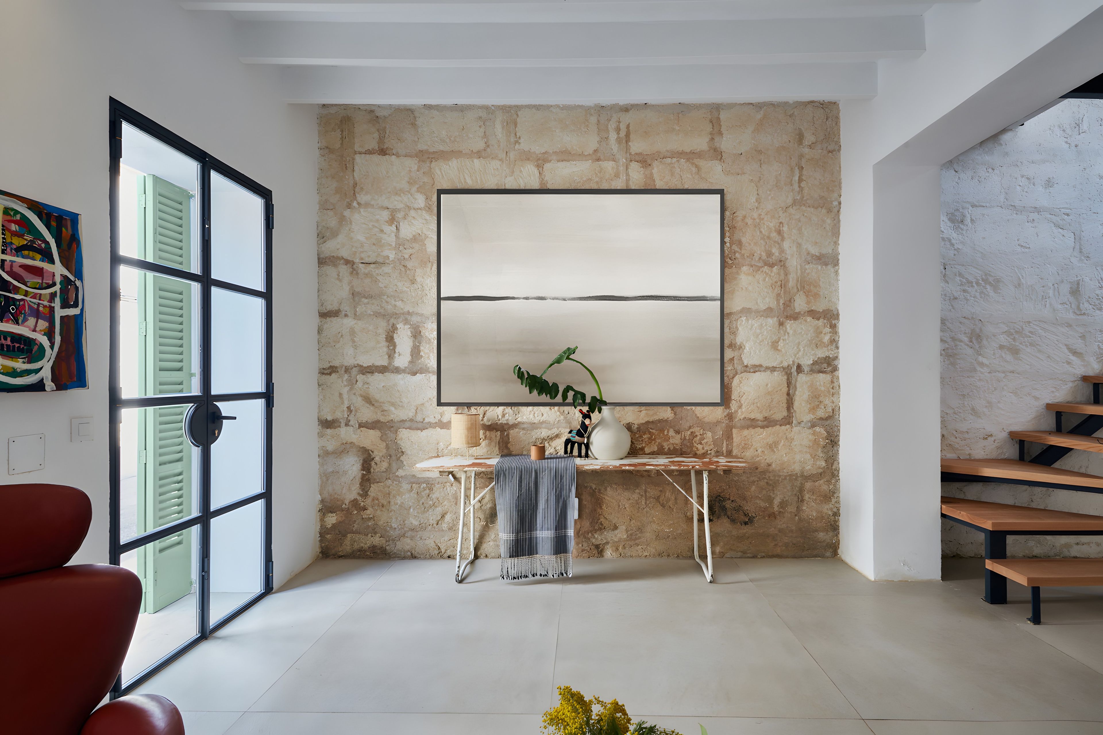 Una habitación con pared de piedra, un cuadro abstracto grande, un escritorio con una planta y una silla roja.