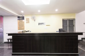 A hotel reception desk with a dark wood finish, a reception sign, a fan, and a window with a metal grill. - Capital O Regal suites (Bangalore)