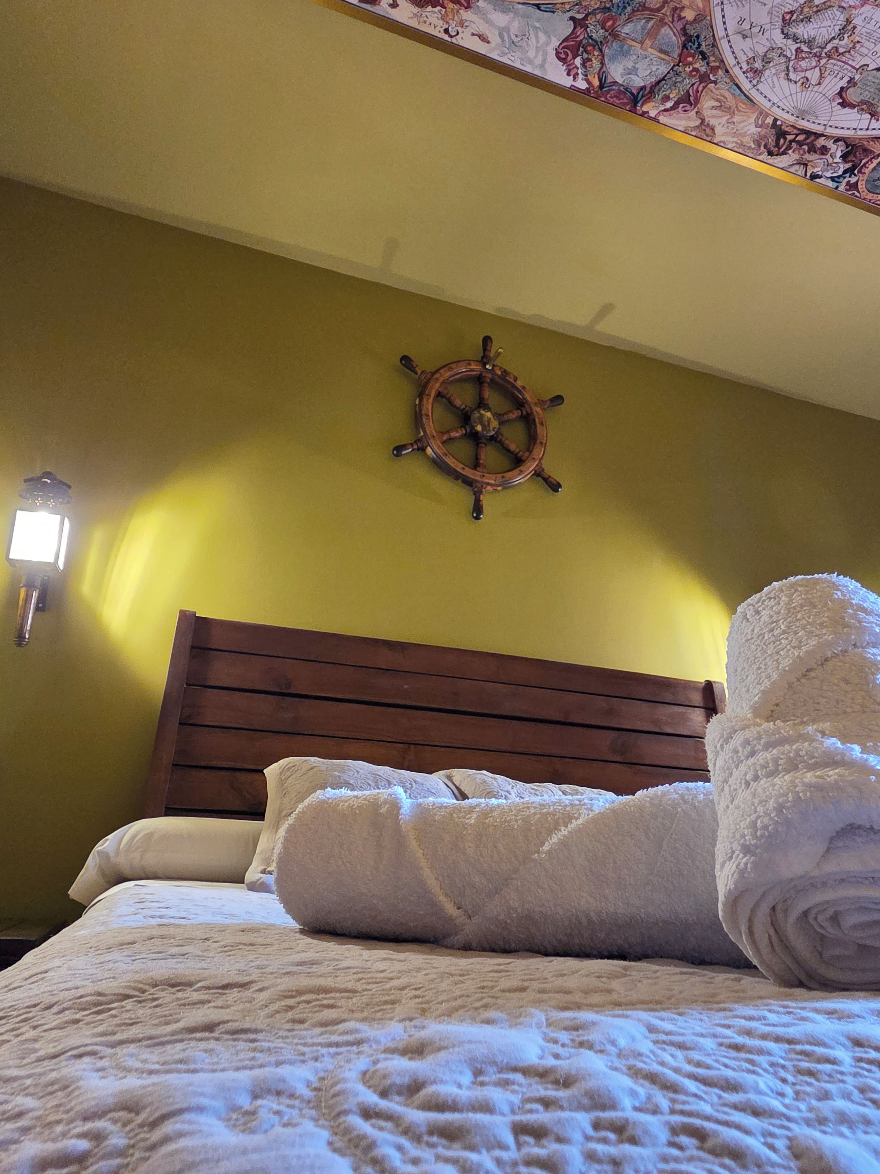 Una cama con cabecera de madera, un timón de barco fijado a la pared y una toalla blanca enrollada.