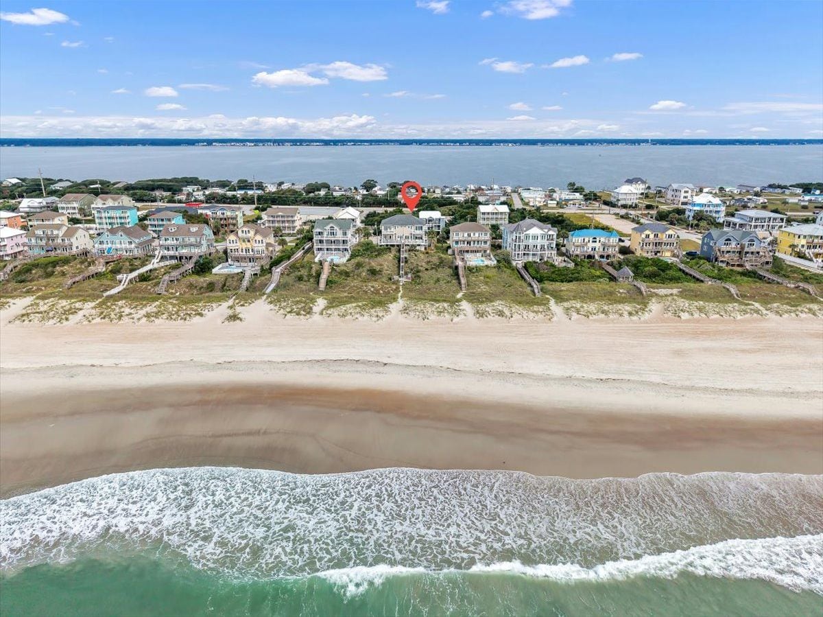 An aerial view showcases the property's prime beachfront location, with the ocean stretching before a row of coastal homes and calm bay waters beyond.