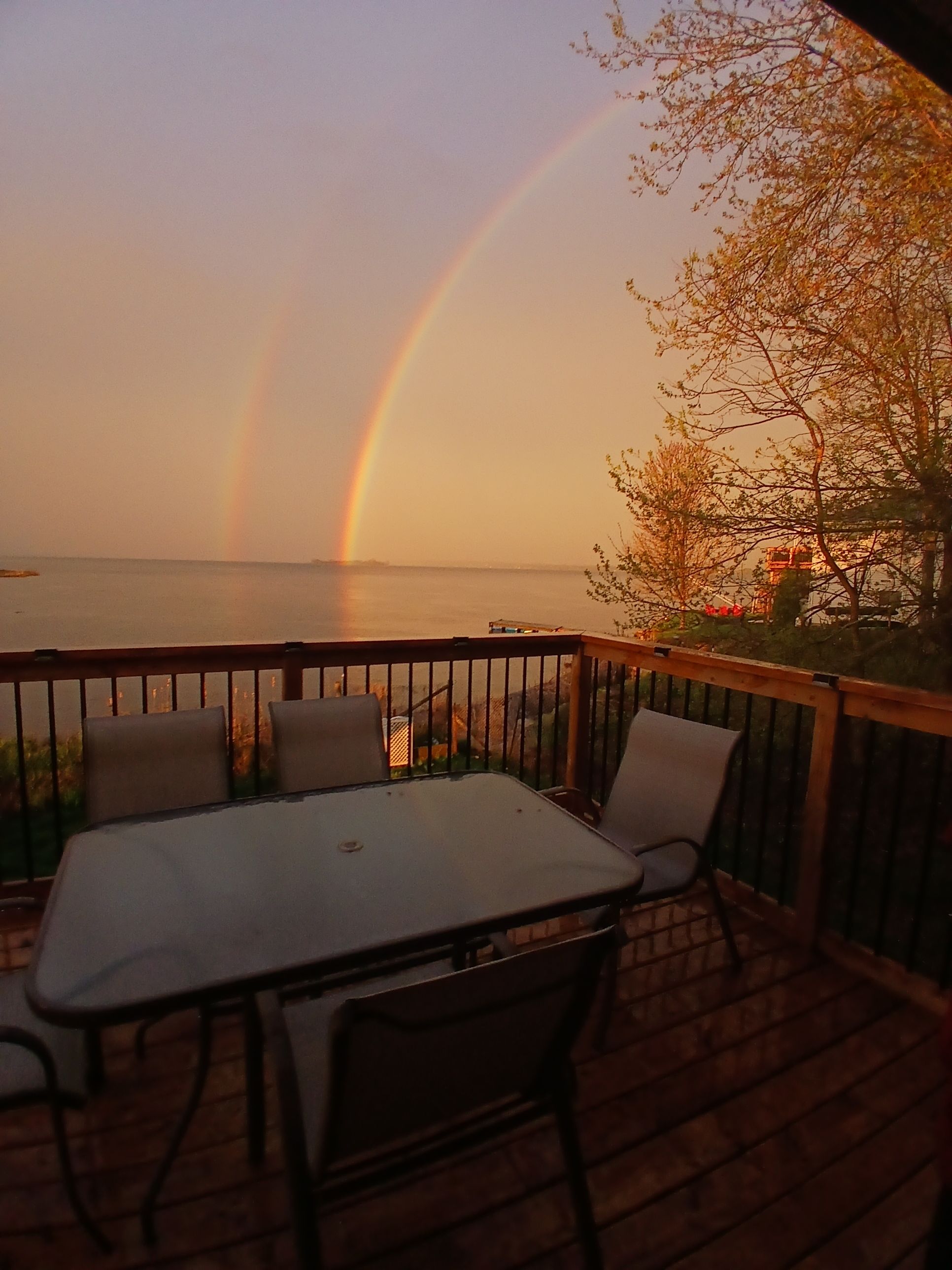 A deck with a table and chairs, overlooking a body of water with a rainbow.
